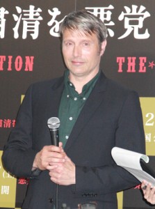 マッツ・ミケルセン両手でマイクを持つ、の図